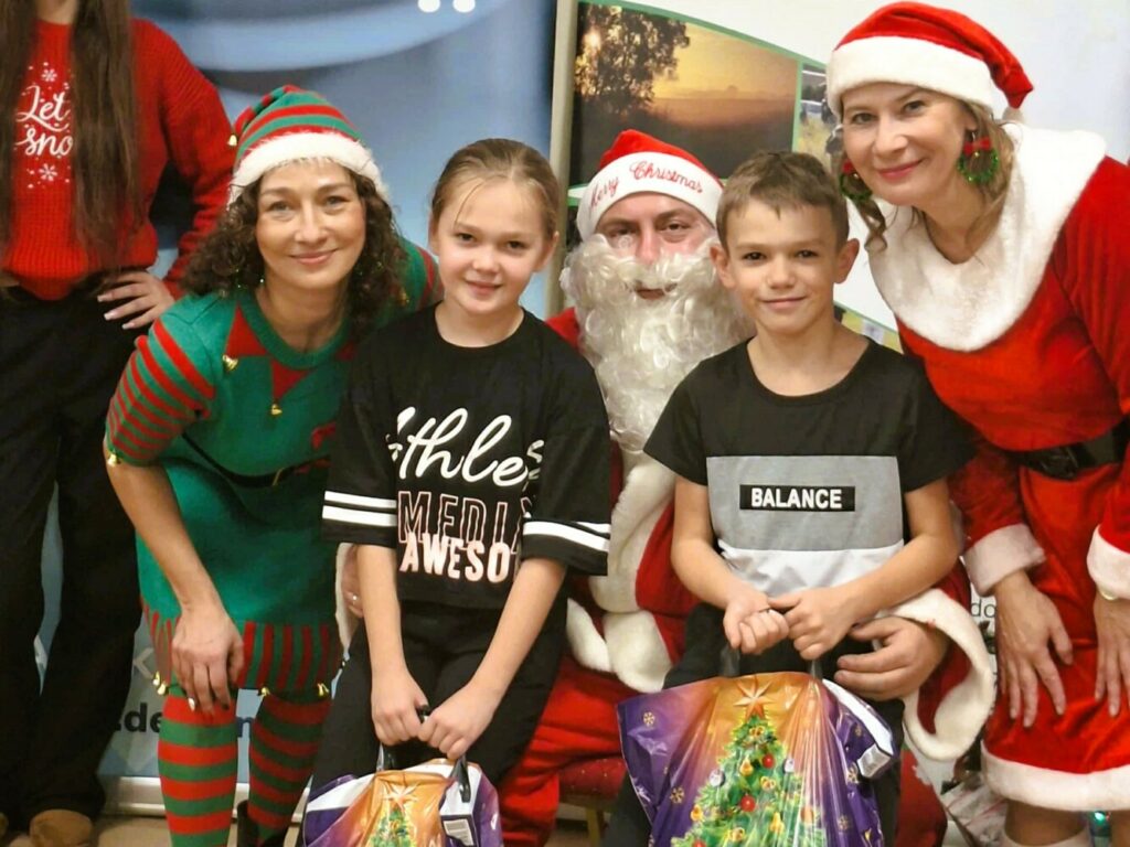 Grupowe zdjęcie z Mikołajek w Dołujach: pośrodku siedzi Święty Mikołaj, a przed nim stoją dwoje uśmiechniętych dzieci trzymających kolorowe paczki świąteczne. Po lewej stronie stoi prowadząca wydarzenie pracownica GCKiB w zielonym stroju elfa, a po prawej Mikołajka ze Stowarzyszenia na rzecz Rozwoju Sołectwa Dołuje–Kościno, ubrana w czerwony strój świąteczny. Wszyscy patrzą w obiektyw i uśmiechają się.”