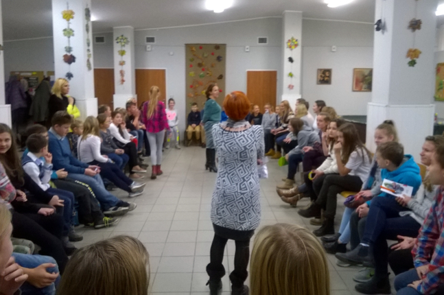 dużo osób siedzących na krzesłach w kręgu i 2 kobiety stojące na środku