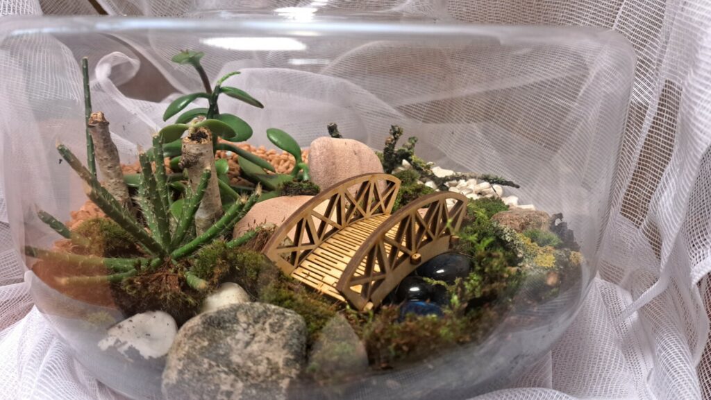 W szklanym naczyniu umieszczone są zielone rośliny,mech poduszkowy, kolorowy piach i kamienie. Udekorowane miniaturowym mostkiem i szklanymi koralami imitującymi wodę.