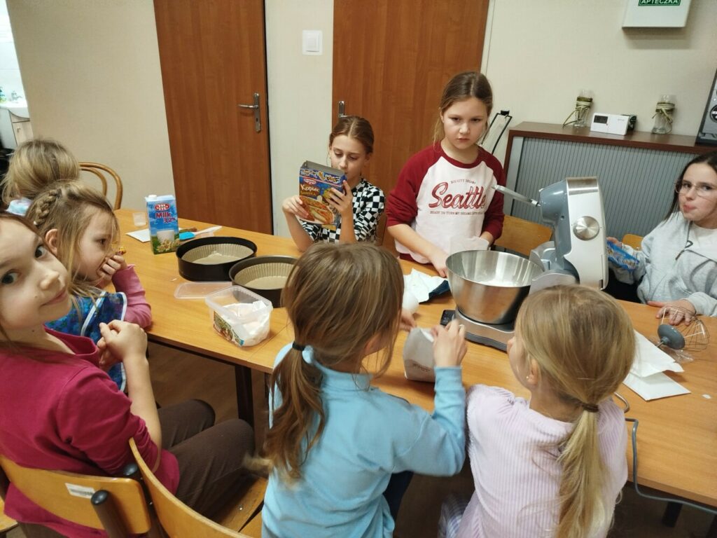 Przy dużym stole w sali świetlicowej siedzi grupa dzieci. Na środku stołu znajduje się mikser kuchenny z metalową miską oraz składniki do pieczenia. Jedno dziecko trzyma opakowanie z przepisem, inne pomagają wsypywać składniki do miski. Dzieci są skupione i zaangażowane w przygotowywanie ciasta