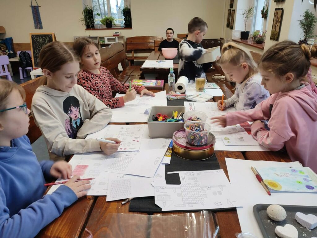 Fotografia przedstawia grupę dzieci siedzących przy stole i malujących kolorowe domki.