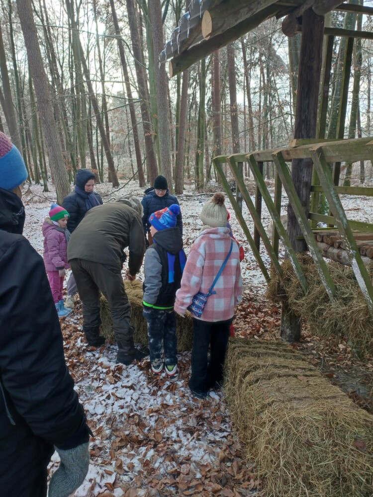 Fotografia przedstawia grupę osób stojących przy paśniku dla zwierzyny leśnej.