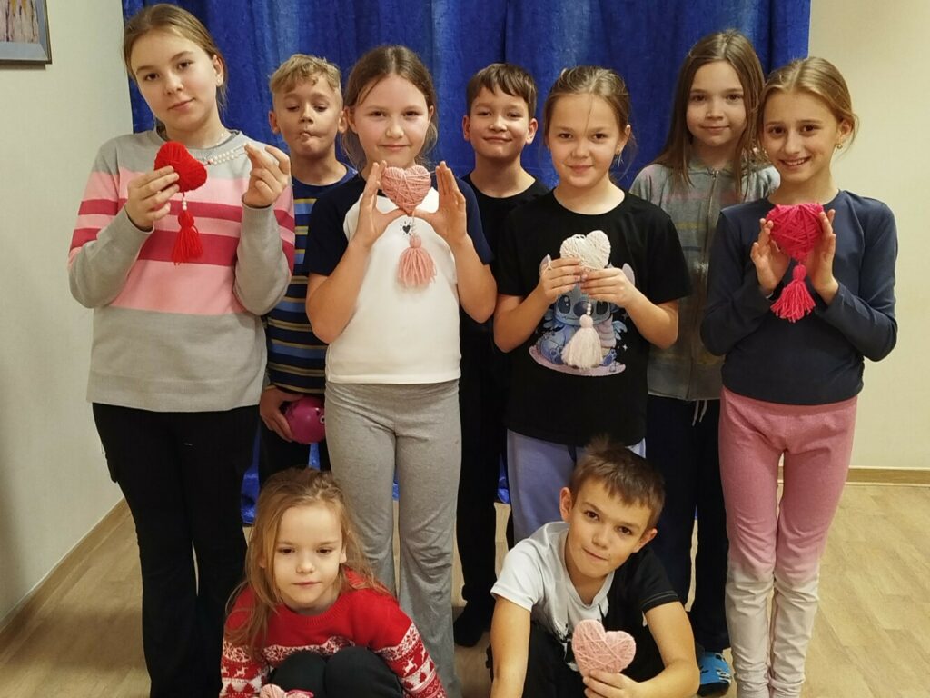 Na zdjęciu widać dziewięcioro dzieci stojących w sali. Każde dziecko trzyma w dłoniach własnoręcznie wykonane serduszko z włóczki z frędzelkiem. Dzieci są uśmiechnięte, ubrane w kolorowe, codzienne stroje. Część dzieci stoi, a dwoje kuca z przodu. Zdjęcie pokazuje radosną atmosferę zajęć plastycznych i wspólnej pracy.