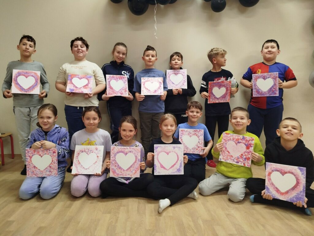 Grupowe zdjęcie dzieci w Klubie GCKiB w Dołujach, stojących i siedzących w dłoniach trzymając własnoręcznie namalowane prace z sercem w odcieniach różowego, czerwonego i fioletu.