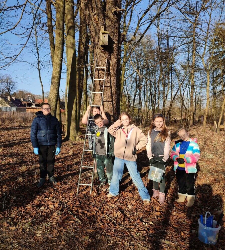 Fotografia przedstawia grupę dzieci stojących przy drzewie, na którym zawieszona jest budka lęgowa dla ptaków.