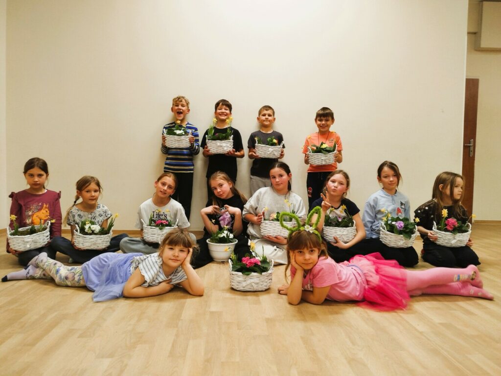 Grupa dzieci podczas warsztatów w Klubie w Dołujach prezentuje własnoręcznie posadzone w koszyczkach wiosenne kwiaty – prymulki, żonkile i hiacynty przygotowane z okazji Dnia Kobiet.