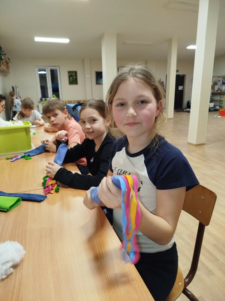 Grupa dzieci siedzi przy stole podczas zajęć plastycznych. Skupione i zaangażowane, własnoręcznie wypełniają kolorowe zabawki dla kotów. Na stole widać materiały, tkaniny. Atmosfera jest radosna i pełna współpracy.