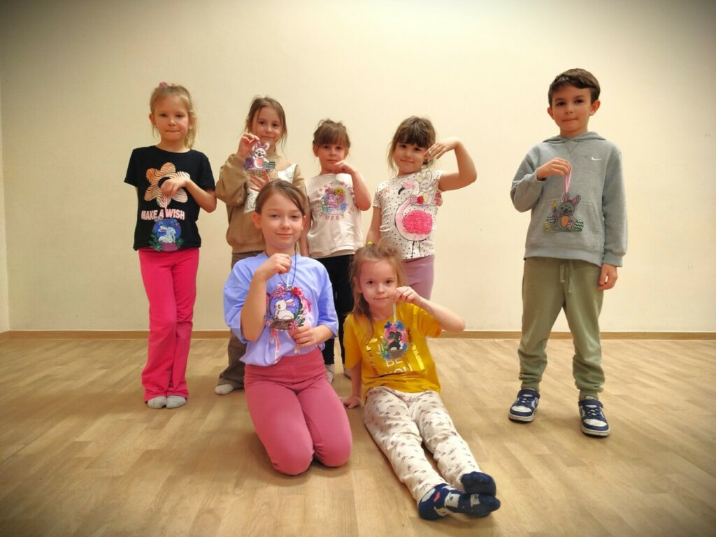 Dzieci stoją pod ścianą w Klubie w Dołujach, prezentując swoje kolorowe, pomalowane wielkanocne łapacze słońca w kształcie króliczków. Widać ich radość i dumę z własnych prac.