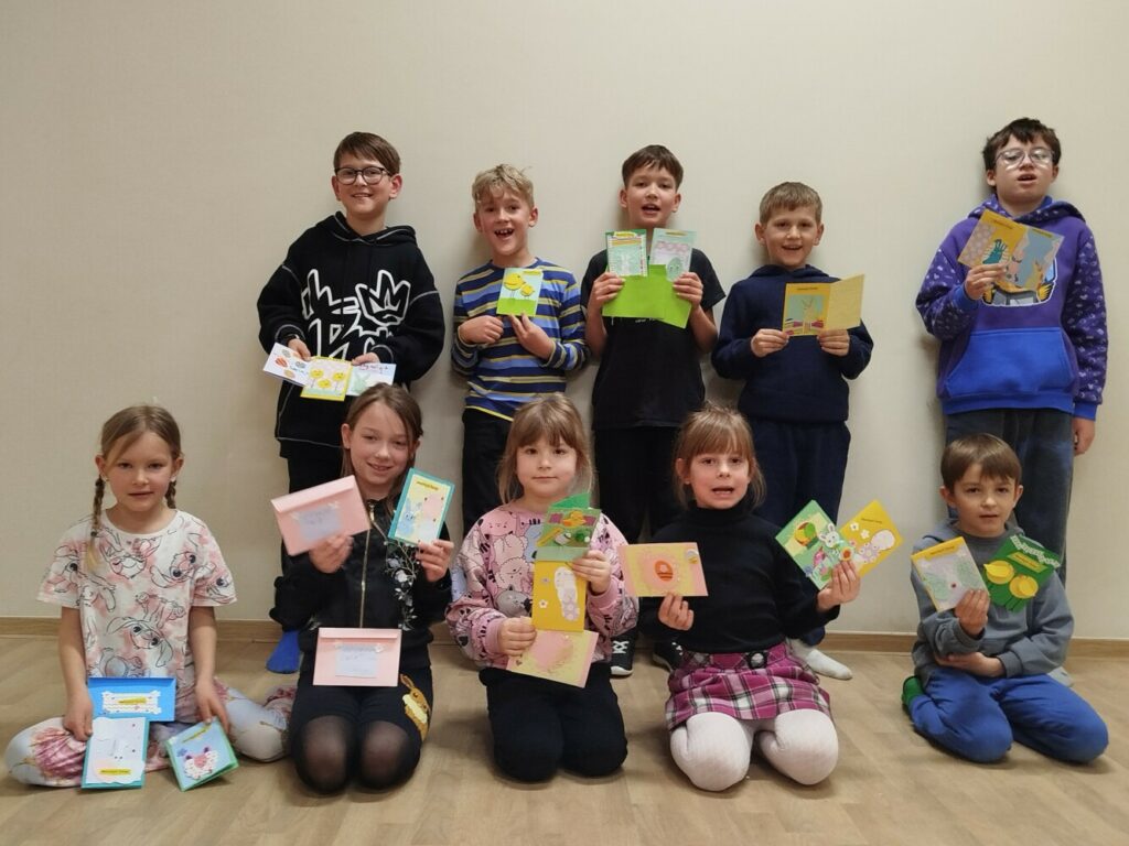 Grupa dzieci w GCKiB w klubie w Dołujach z ręcznie wykonanymi kolorowymi kartkami Wielkanocnymi. Dzieci uśmiechają się do aparatu, widać radość i zaangażowanie.