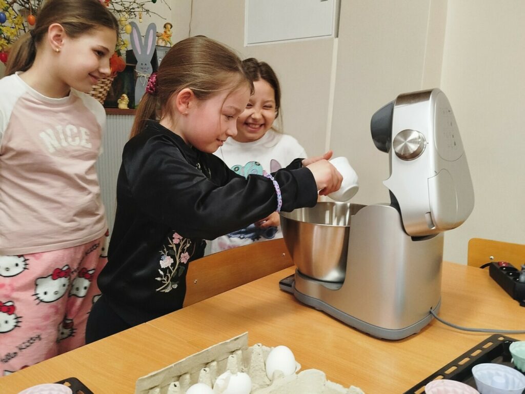 Troje dzieci stoi przy stole i wspólnie przygotowuje ciasto w robocie kuchennym. Jedna z dziewczynek wsypuje składniki do metalowej misy miksera, a pozostałe obserwują z uśmiechem. Na stole obok leżą jajka i akcesoria do pieczenia. Zdjęcie zostało wykonane podczas warsztatów kulinarnych dla dzieci w Klubie w Dołujach.