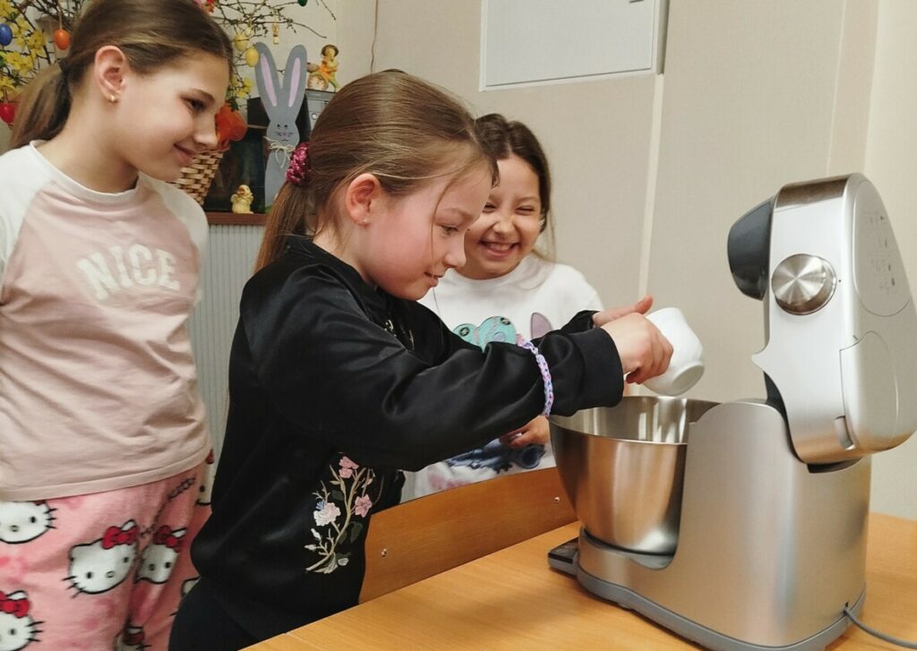 Troje dzieci stoi przy stole i wspólnie przygotowuje ciasto w robocie kuchennym. Jedna z dziewczynek wsypuje składniki do metalowej misy miksera, a pozostałe obserwują z uśmiechem. Na stole obok leżą jajka i akcesoria do pieczenia. Zdjęcie zostało wykonane podczas warsztatów kulinarnych dla dzieci w Klubie w Dołujach.