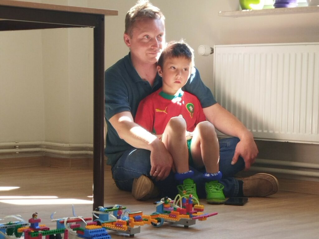Tata z synkiem siedzący na podłodze podczas zajęć z Robotyki . W prawym rogu Kolorowe roboty lego gotowe do rozpoczęcia meczu piłki.