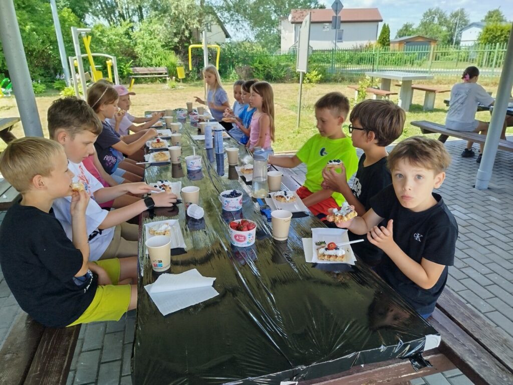 Grupa dzieci siedzi przy długim stole pod zadaszeniem i je gofry z bitą śmietaną oraz owocami. Na stole stoją kubki, owoce i dodatki, a wokół panuje letnia, radosna atmosfera wspólnego posiłku.