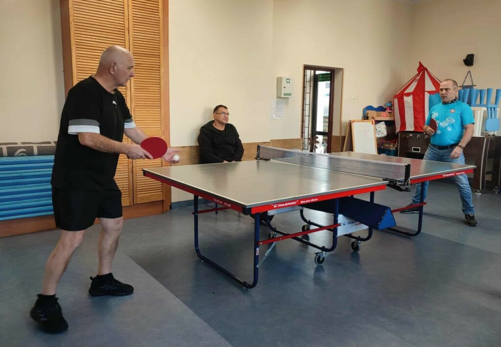 Fotografia przedstawia mężczyzn grających przy stole w tenisa stołowego.
