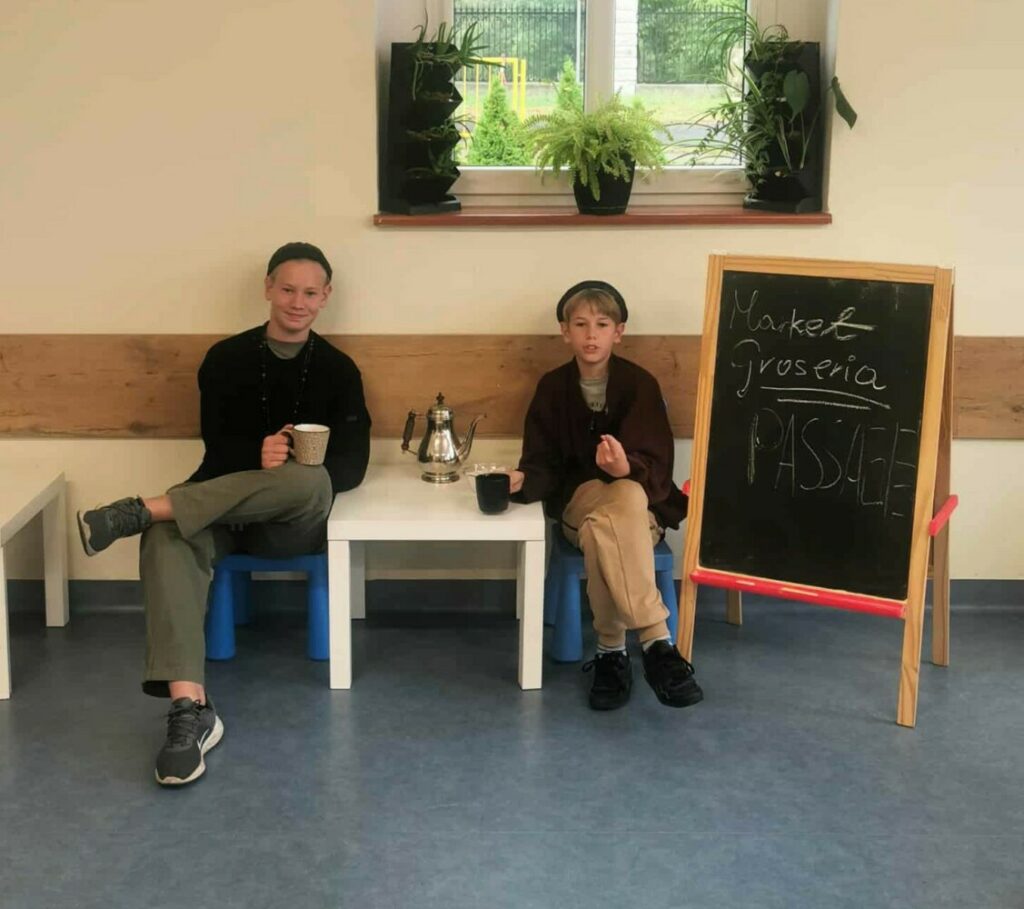 Fotografia przedstawia dwóch siedzących chłopców. Obok nich mała czarna tablica z napisem: "Market Groseria".