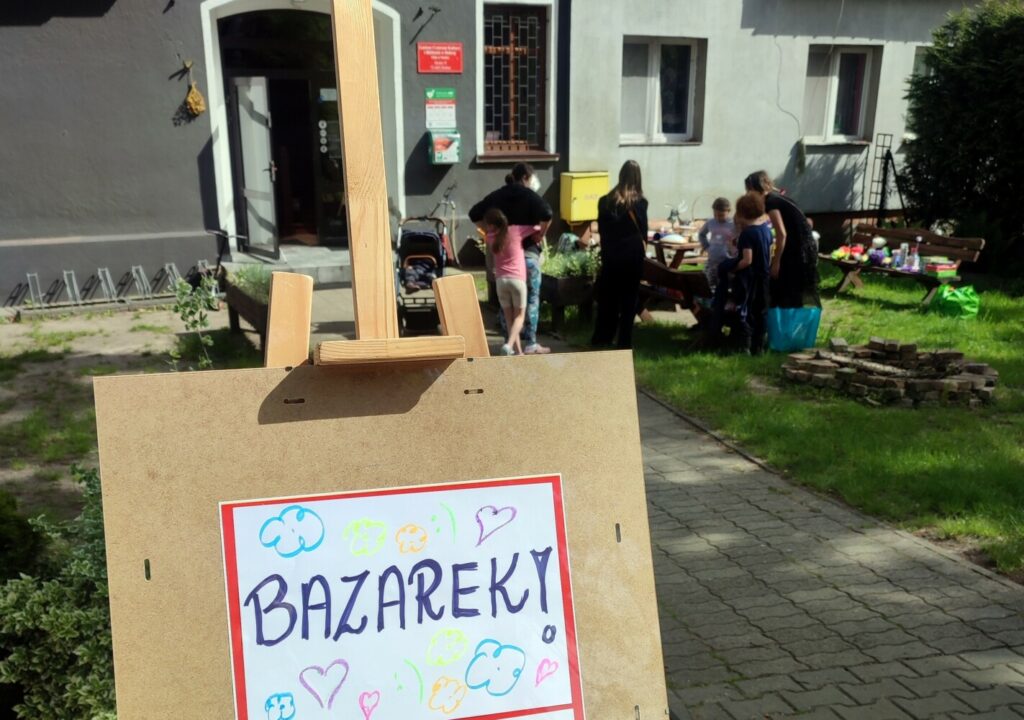 Fotografia przedstawia sztalugę z widocznym napisem "Bazarek". W oddali grupa ludzi przeglądająca różne przedmioty.