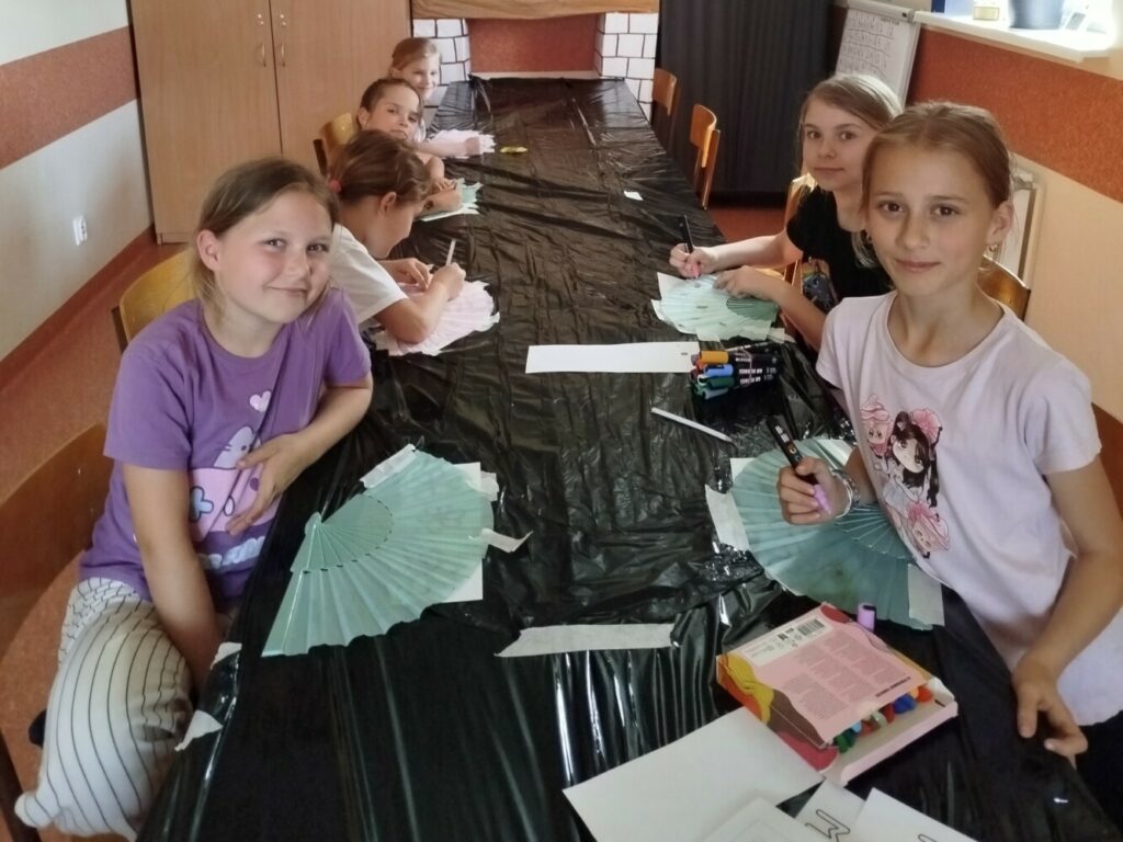 Na zdjęciu widać grupę dziewczynek siedzących przy długim stole. Dzieci biorą udział w warsztatach – skupione dekorują różne przedmioty, m.in. wachlarze i breloczki, które mają posłużyć jako prezenty z okazji Dnia Matki. Każde dziecko ma przed sobą materiały plastyczne – flamastry, pisaki, elementy do ozdabiania.