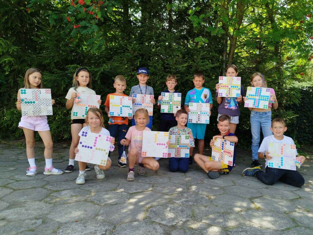 Grupa kilkunastu dzieci stoi na świeżym powietrzu, uśmiechając się do zdjęcia. Każde z nich trzyma w rękach własnoręcznie wykononą pracę plastyczną przedstawiającą kolorowe plansze do gry „Chińczyk”.