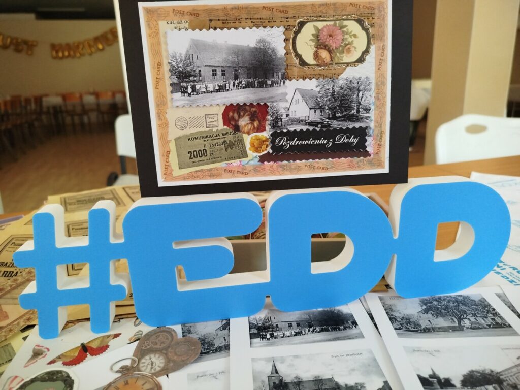 Stół z materiałami do scrapbookingu: kolorowe papiery, stare fotografie Dołuj i Kościna oraz duży, niebieski napis #EDD. Zdjęcie wykonane podczas warsztatów ‘Miejsca ludzkich wspomnień’ w Klubie w Dołujach.