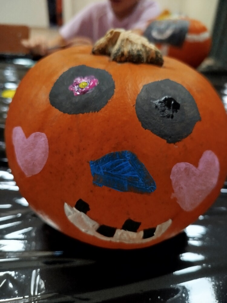 Pomarańczowa dynia z namalowanymi czarnymi oczami, różowymi serduszkami na policzkach, niebieskim nosem i szerokim uśmiechem.