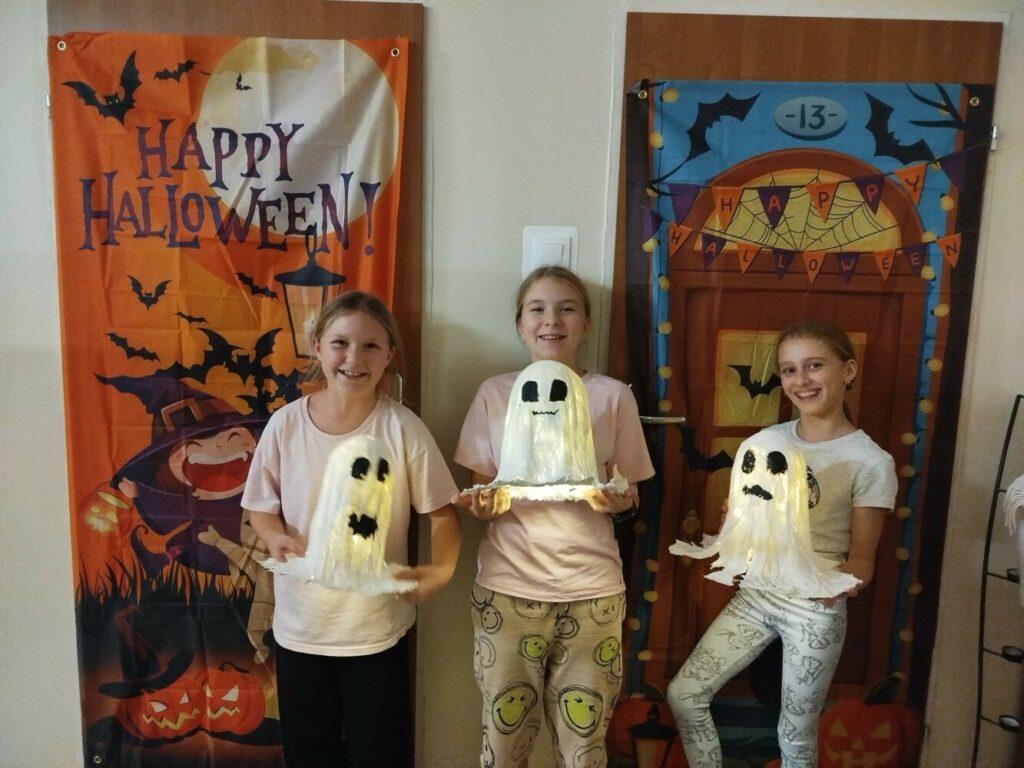 Trzy uśmiechnięte dziewczynki stoją przed kolorowymi halloweenowymi dekoracjami. Każda trzyma w rękach samodzielnie wykonanego duszka z bandaży, z namalowaną buźką i delikatnym, świetlistym wnętrzem.
