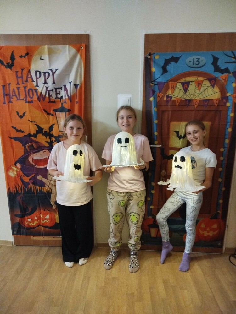 Trzy uśmiechnięte dziewczynki stoją przed kolorowymi halloweenowymi dekoracjami. Każda trzyma w rękach samodzielnie wykonanego duszka z bandaży, z namalowaną buźką i delikatnym, świetlistym wnętrzem.