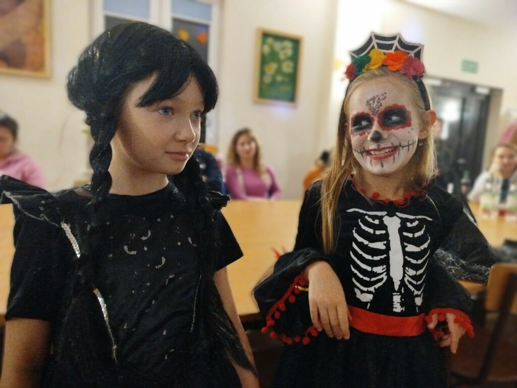 Dwie dziewczynki w halloweenowych przebraniach stoją obok siebie w sali. Jedna ma długie, ciemne warkocze i czarny strój przypominający postać Wednesday Addams. Druga ma pomalowaną twarz w stylu meksykańskiego Dnia Zmarłych i ubrana jest w czarną sukienkę z nadrukiem szkieletu oraz kolorową opaskę z kwiatami i pajęczyną. Obie uśmiechają się, w tle widać siedzące osoby.