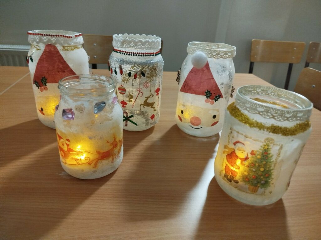 Na zdjęciu stoi kilka szklanych słoików ozdobionych techniką decoupage. Każdy z nich ma świąteczne wzory: renifery, bombki, choinki i Mikołaje. Słoiki pokryte są brokatem i delikatnymi kolorami farb, a wewnątrz kilku z nich widać ciepłe światełka, które tworzą przytulny, zimowy nastrój. Dekoracje ustawione są na stole, a tło jest lekko rozmazane, dzięki czemu lampiony przyciągają uwagę.