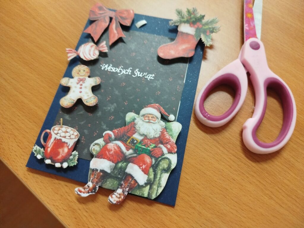 Kartka świąteczna wykonana przez dziecko. Na granatowym tle znajdują się naklejone dekoracje: pierniczek, cukierek, kokarda, świąteczna skarpeta, kubek gorącej czekolady oraz duża ilustracja Mikołaja siedzącego w fotelu.