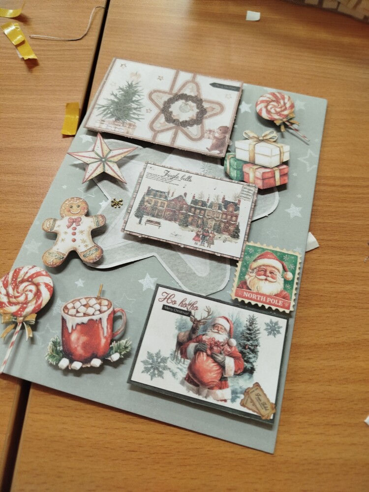 Na zdjęciu znajduje się ręcznie wykonana kartka świąteczna ozdobiona elementami w stylu retro: są obrazki z Mikołajem, choinką, pierniczkiem, gorącą czekoladą, lollipopami i prezentami. Kartka leży na drewnianym blacie, a całość utrzymana jest w zimowych kolorach z akcentami czerwieni i zieleni.