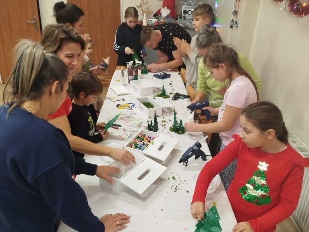 Uczestnicy Rodzinnych Warsztatów Świątecznych w Klubie GCKiB w Dołujach stoją przy stołach i wybierają ozdoby oraz dekoracje do świątecznych stroików z drewnianych choinek.