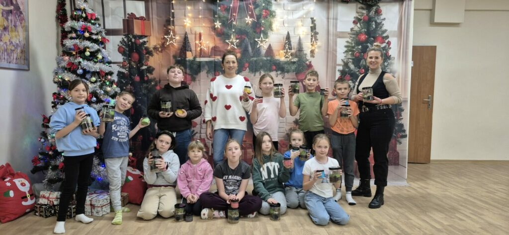 Grupa dzieci i młodzieży wraz z dwiema prowadzącymi stoi i siedzi w sali Klubu w Dołujach, na tle świątecznej dekoracji z choinkami i lampkami. Każde dziecko trzyma w dłoniach słoik z własnoręcznie wykonanym lasem w słoiku lub naturalną solą do kąpieli, przygotowanymi podczas warsztatów ekologicznych.
