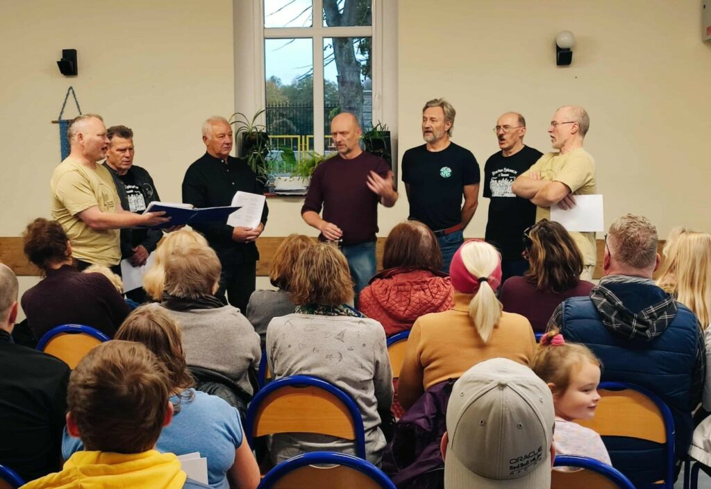 Fotografia przedstawia śpiewających mężczyzn, przed którymi siedzi publiczność.
